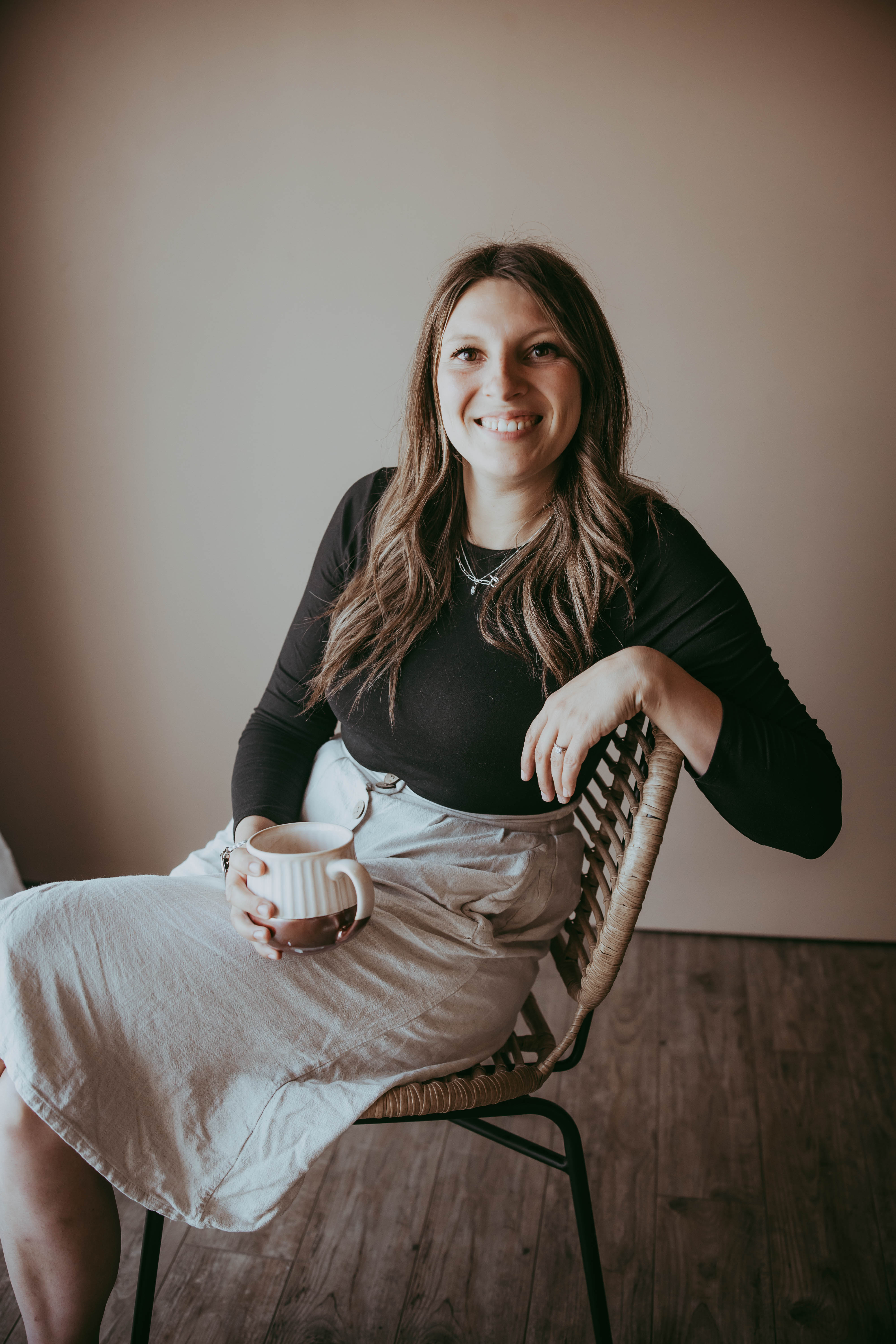 Smiling woman with long brown hair sitting in a wicker chair, holding a ceramic mug and wearing a black top and light gray skirt.